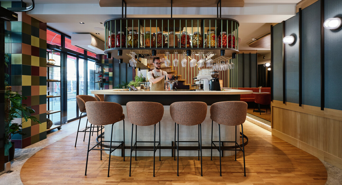 Ein stilvoller Barkeeper in einem modernen Café des Henriette Stadthotels, umgeben von eleganten Stühlen und hängenden Weingläsern.