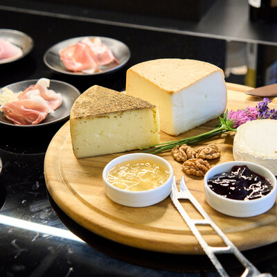 A cheese platter with assorted cheeses, nuts, honey, jam, and lavender on a wooden board. Plates of cured meats are in the background.