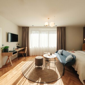 A cozy, sunlit suite at City hotel Henriette features a king bed, blue sofa, round rug, desk, and smart TV in warm, modern decor.