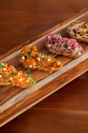 A wooden platter with gourmet open-faced sandwiches, topped with various meats, vegetables, and garnishes.