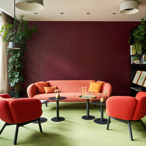 Modern lounge area with red seating, green accents, and plants, creating a cozy and stylish atmosphere. Large windows bring in natural light.