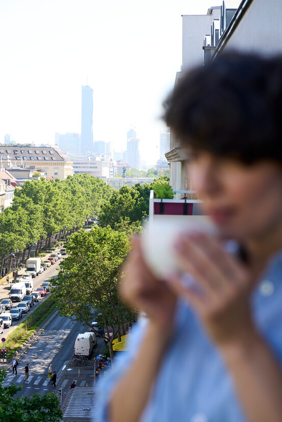 View of Vienna street from a balcony with blurred person holding a cup, trees lining the road, and modern buildings in the background.
