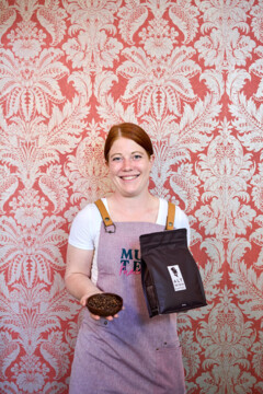 A woman in an apron holding coffee beans and a bag of Alt Wien Kaffee, standing against a decorative red wallpaper.