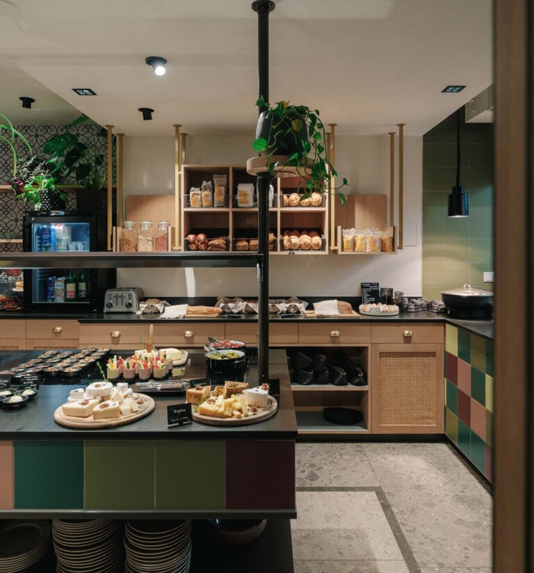 Hotel Henriette breakfast buffet featuring fresh breads, cheeses, fruit, and cereals in a warm, plant-filled dining area.