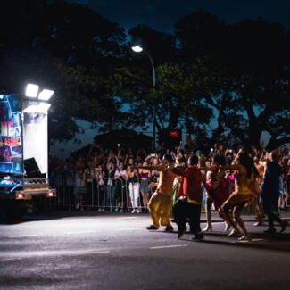 Colorful night parade with dancers performing in vibrant costumes as a cheering crowd lines the street.