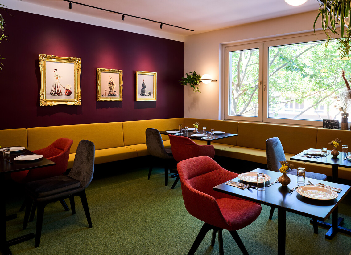 Cozy hotel dining area with colorful chairs, elegant wall art, and a large window overlooking greenery.