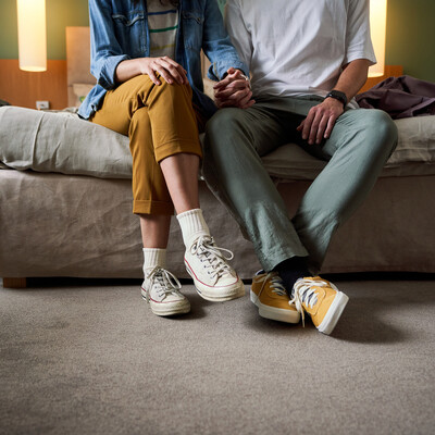 A couple sits on a hotel bed, holding hands, with a suitcase nearby.