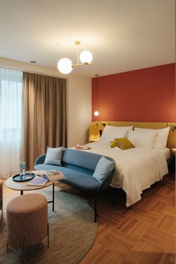 Stylish Hotel Henriette room with plush bed, coral accent wall, blue settee, round table with water and books, warm lights, and parquet floor.