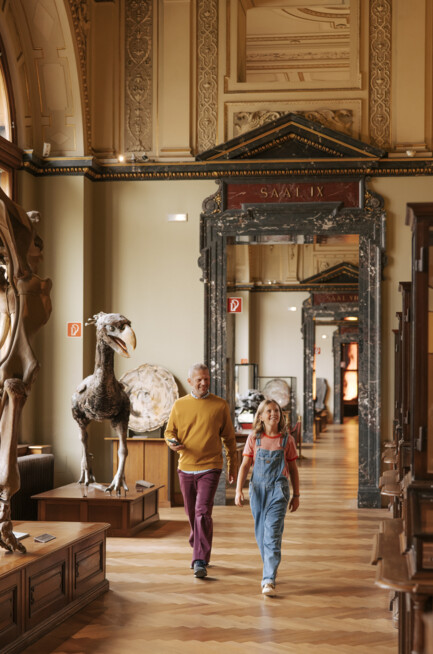 Father and daughter walking through a hall with dinosaurs of the Natural History Museum Vienna – a holiday outing with Henriette's Vienna Explorer Package.