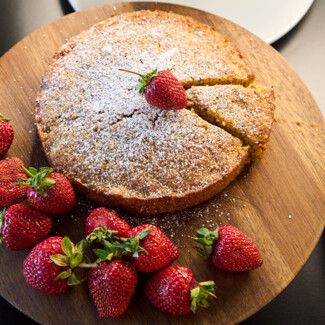 A wooden platter with a sliced cake dusted with powdered sugar, surrounded by fresh strawberries. Donuts and another cake are in the background.