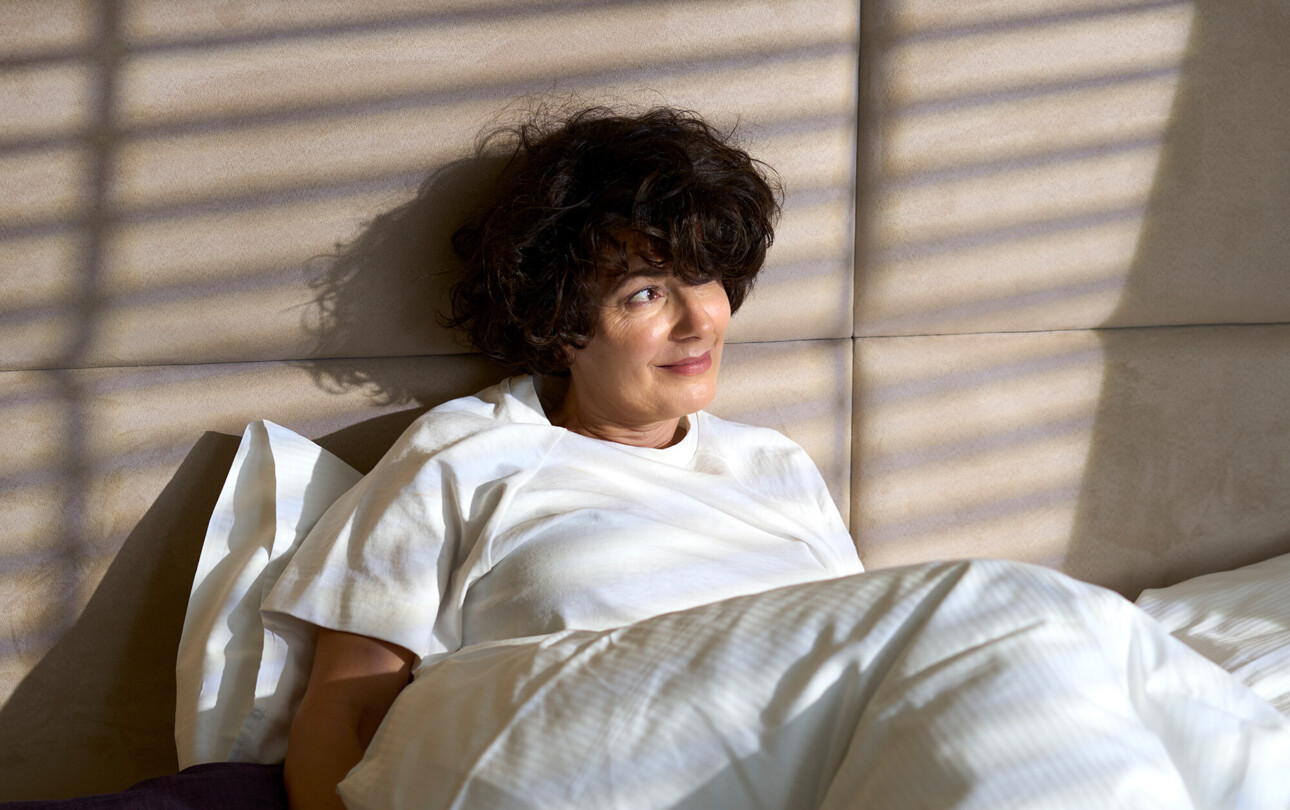 Eine Frau entspannt im Bett, im Henriette Stadthotel, mit Sonnenlicht durch Jalousien.