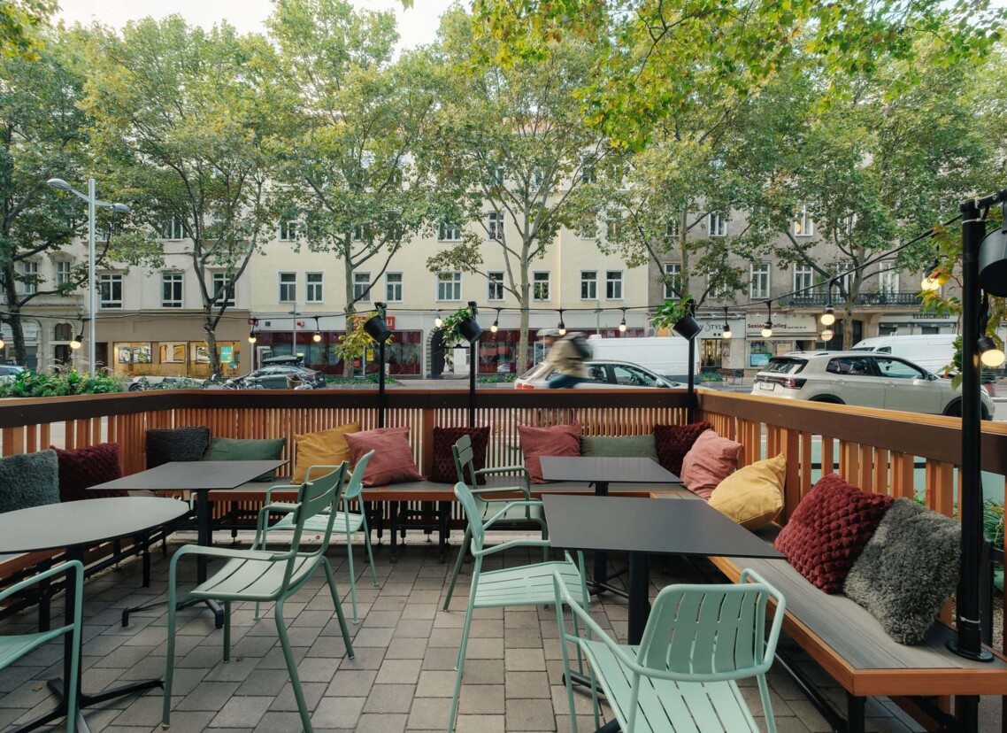 Gemütliche Terrasse der Bar "der schöne Ernst" mit Tischen, grünen Stühlen und bunten Kissen unter Bäumen an der Straße.