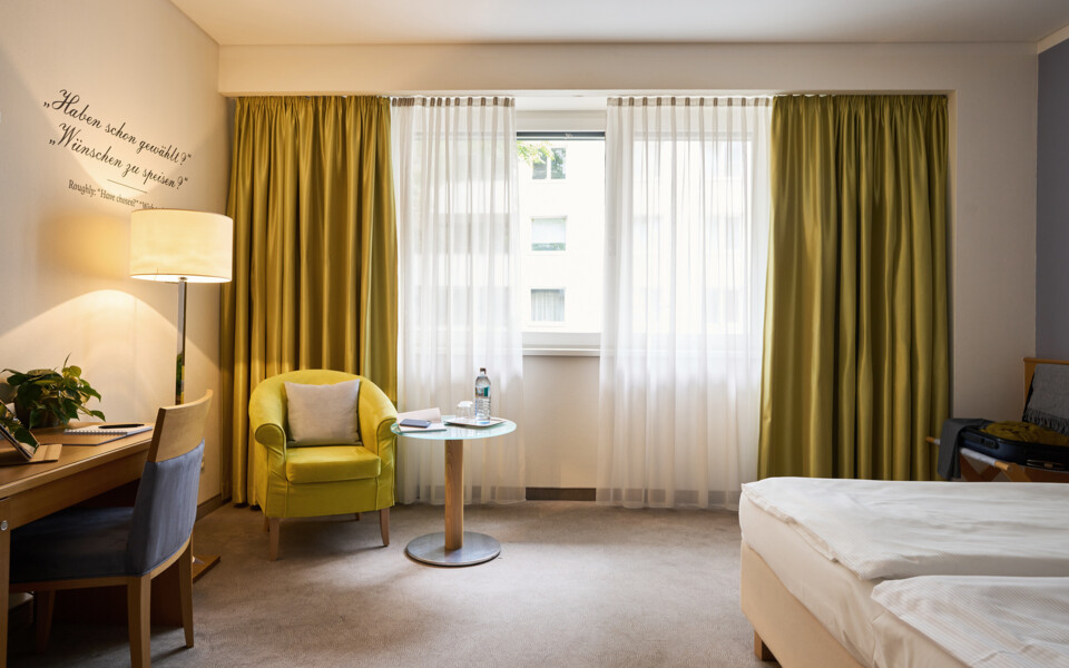 Gemütliches Hotelzimmer im Henriette Stadthotel mit grünem Sessel, Schreibtisch und Fenster mit Vorhängen.