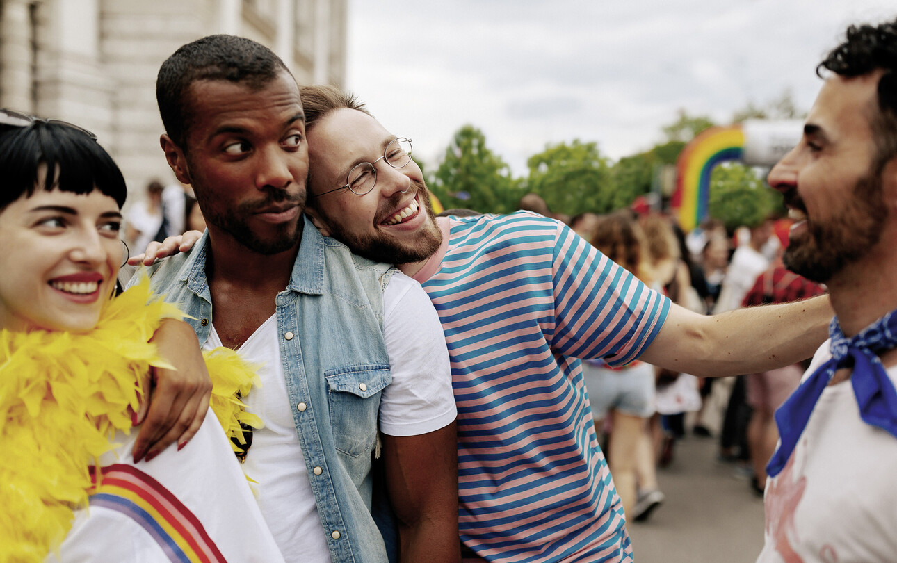 Eine Gruppe fröhlicher Menschen feiert draußen, mit Regenbogenfarben und festlicher Kleidung.