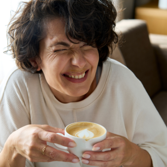 Woman with short curly hair holding a cup of coffee with milk foam smiling with closed eyes sitting on a sofa.