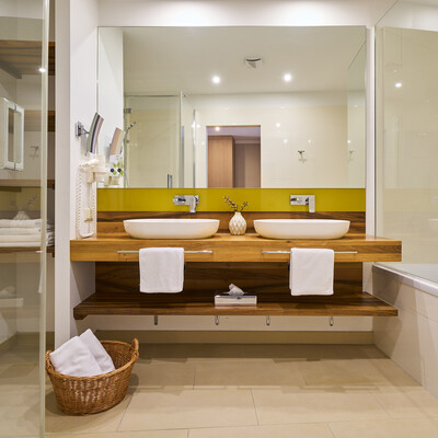 Modern bathroom with double sinks, wooden countertop, and large mirror. Towels are neatly arranged, and a wicker basket sits below.