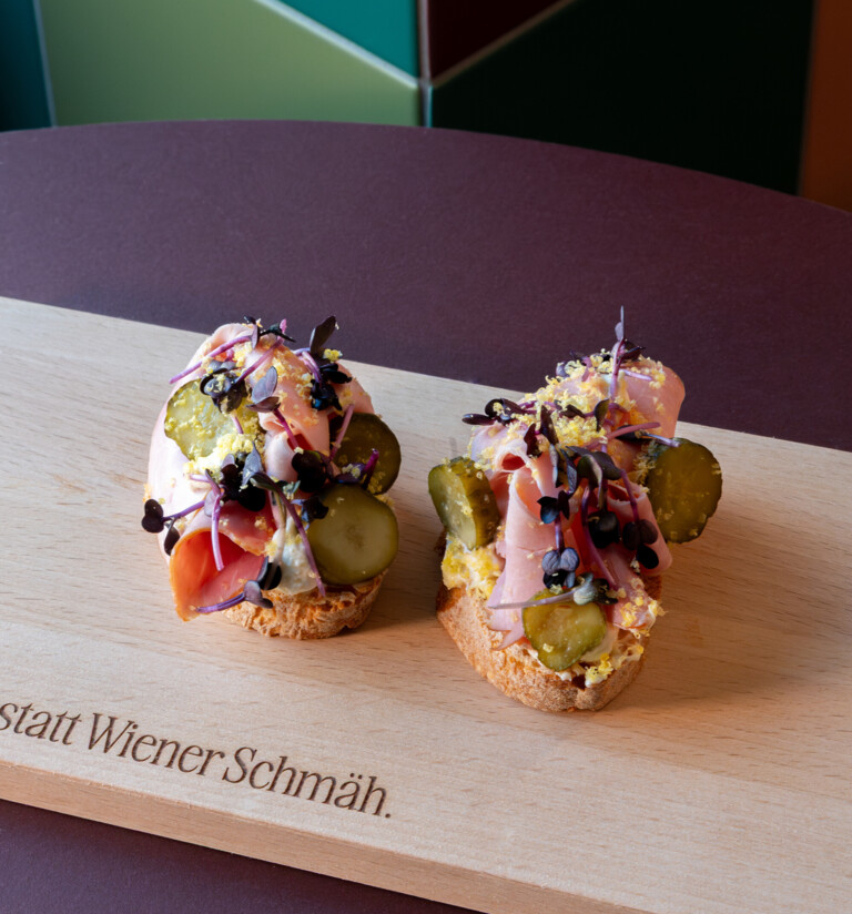 Two open-faced sandwiches topped with ham, pickles, egg spread and microgreens on a wooden board, served at Der schöne Ernst.