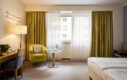 Gemütliches Hotelzimmer im Henriette Stadthotel mit grünem Sessel, Schreibtisch und Fenster mit Vorhängen.