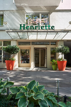Entrance of Hotel Henriette with green signage, potted plants, and glass doors, surrounded by lush greenery.