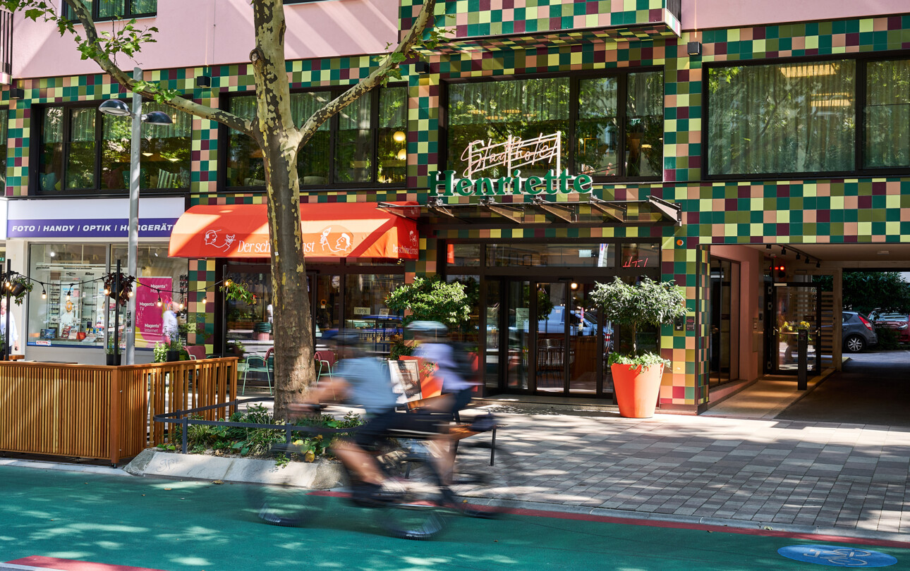Eingang des 4* Stadthotel Henriette in Wien mit bunter Kachel-Fassade, Glasportal und Radweg; gemütliches Straßencafé daneben.