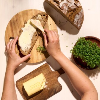 Hands spreading butter on a slice of organic bread on a round wooden board next to a wooden board with a block of organic butter and a small pot of fresh garden cress.