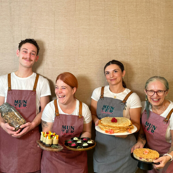 Five smiling people wearing aprons, holding various baked goods and a book.