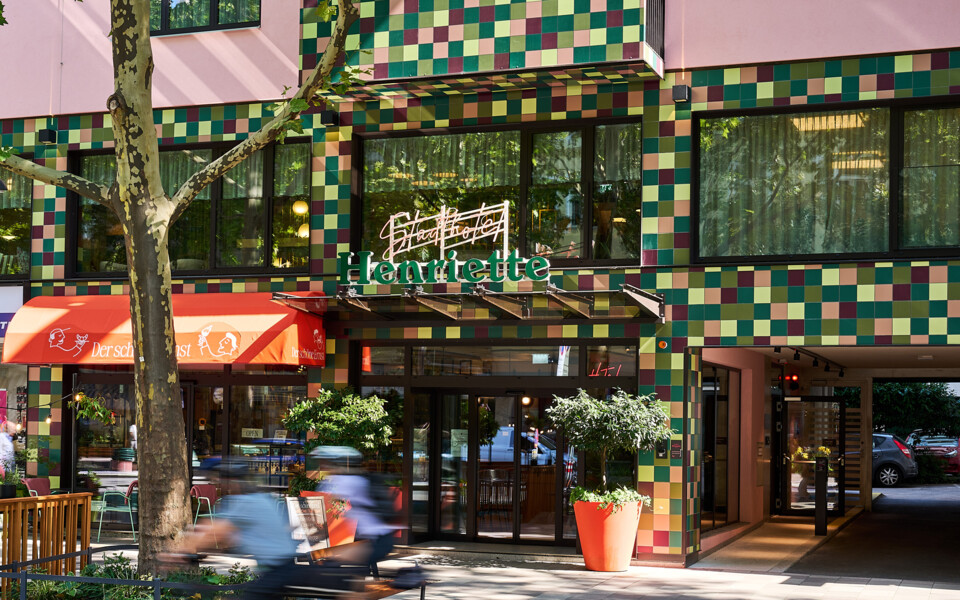 Colorful facade of City hotel Henriette in Vienna, showing the entrance, cafe terrace, and a cyclist passing on the green bike lane.