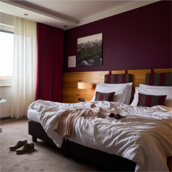 Spacious standard room with a double bed, white organic cotton bed linen, decorative cushions with a red and grey striped pattern, a wooden headboard, a dark red wall, and a window with white and red curtains.