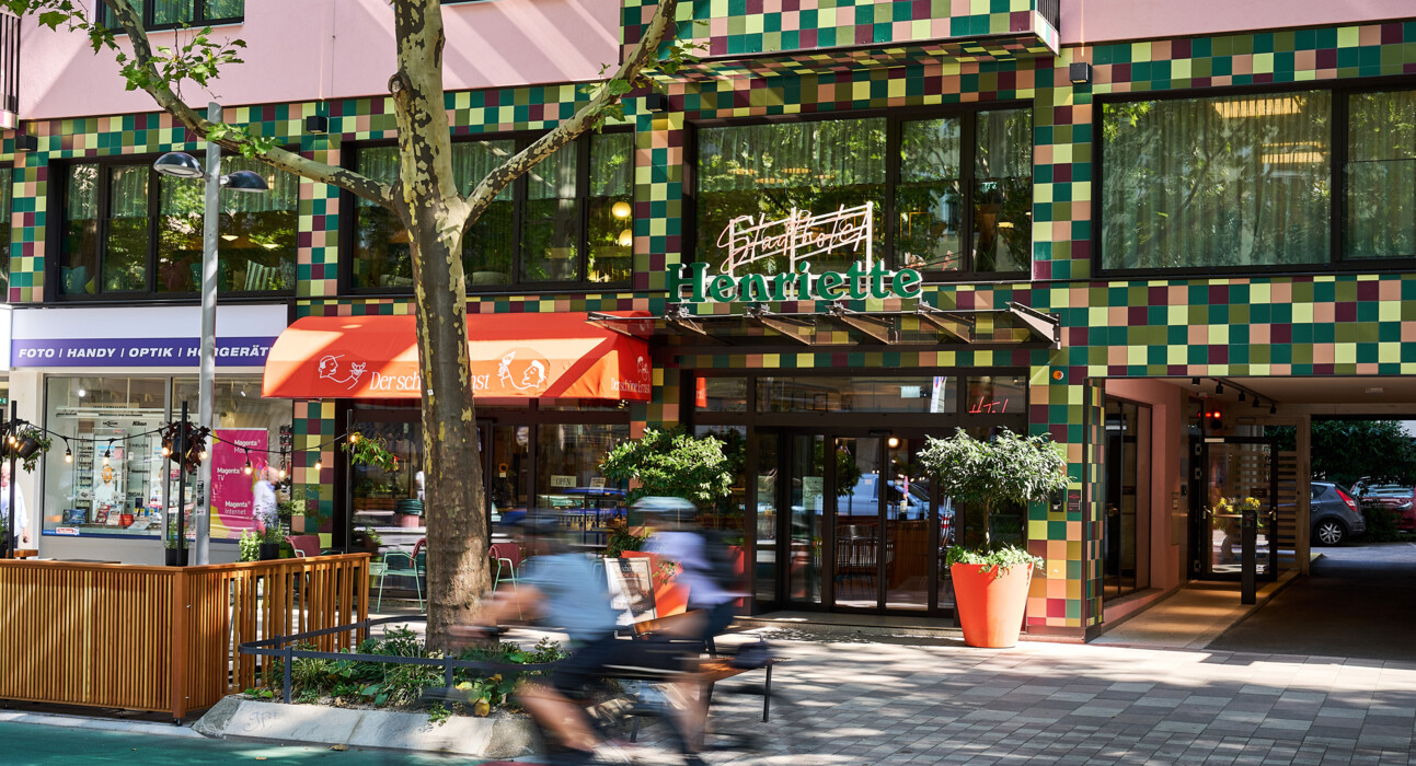 Eingang des 4* Stadthotel Henriette in Wien mit bunter Kachel-Fassade, Glasportal und Radweg; gemütliches Straßencafé daneben.
