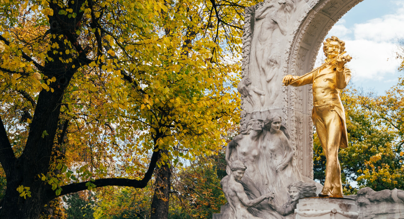Gold-colored bronze statue of Johann Strauss in front of a white stone arch with reliefs in Vienna Stadtpark, surrounded by autumn yellow trees.