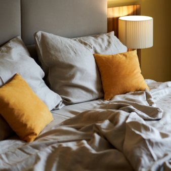 Gemütliches Bett im Hotel Henriette in Wien, mit Bio-Bettwäsche, zwei großen Kissen und zwei kleinen senffarbenen Kissen neben einer weißen Nachttischlampe.